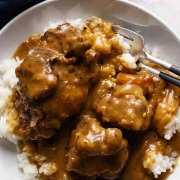 A white plate filled with steamed white rice topped with chunks of meat and gravy. The dish is served with black chopsticks and a fork placed on the side. The meal looks savory, and the gravy appears to be rich and thick. A blue napkin is partially visible to the left.