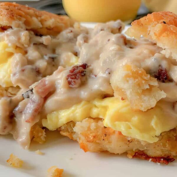 The image shows a biscuit sandwich filled with scrambled eggs, topped with creamy gravy and bacon pieces, all arranged on a white plate.
