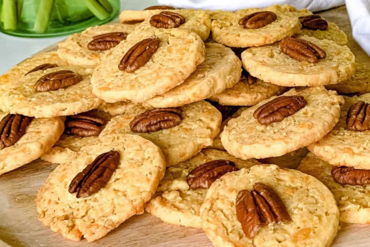 A pile of freshly baked cookies topped with pecan halves on a wooden board. The cookies are golden brown and have a textured surface. In the background, there is a blurred view of a green plant.