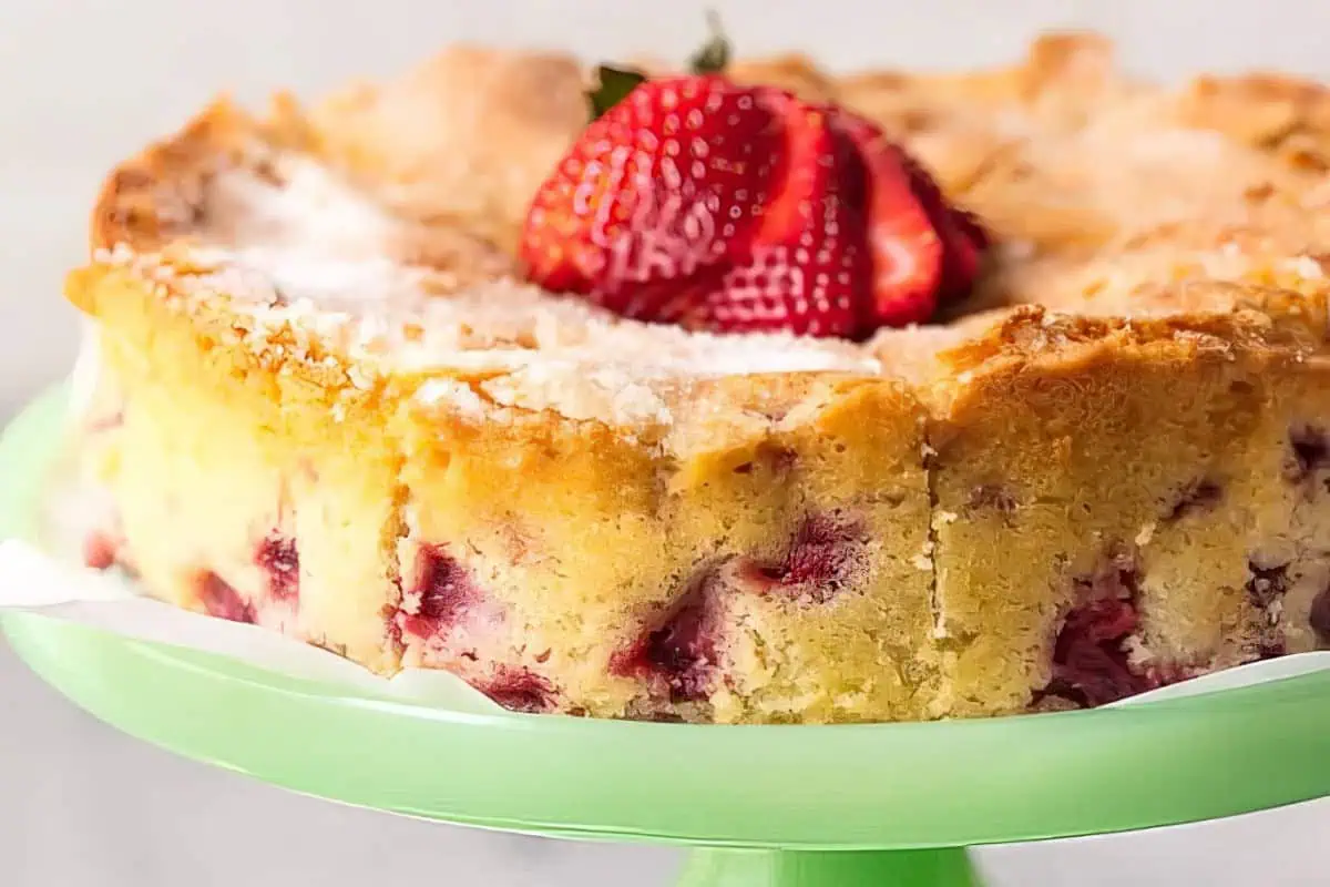 A close-up of a strawberry cake topped with a halved fresh strawberry. The cake has a golden crispy crust on top and visible strawberry pieces inside. It is placed on a green cake stand with a white paper underneath.