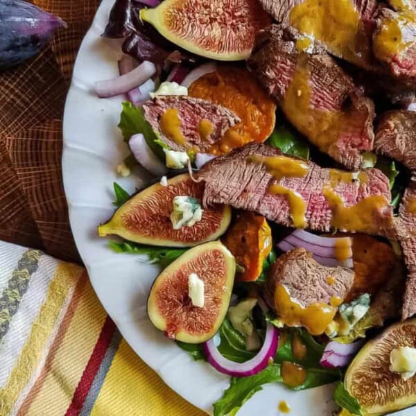 A plate of steak salad with figs, sweet potatoes, red onions, blue cheese, and greens. A whole fig and striped napkin nearby.