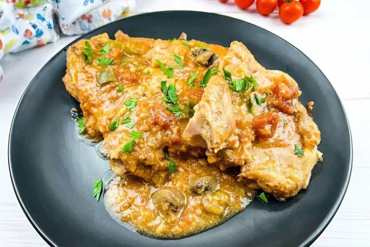 A savory dish of chicken in a rich tomato-based sauce is served on a black plate. The chicken is garnished with fresh parsley and mixed with vegetables like bell peppers and mushrooms. In the background, there are cherry tomatoes and a colorful napkin.