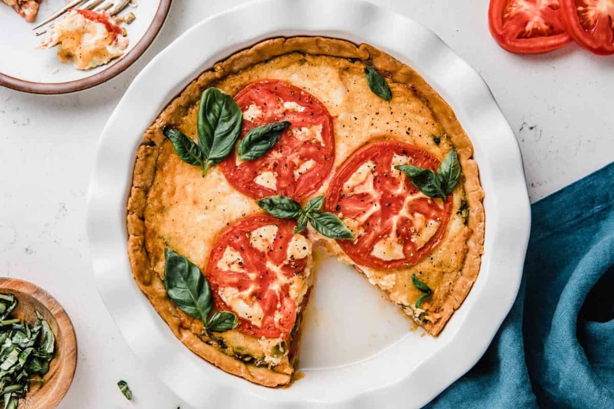 A savory pie with a golden-brown crust is topped with large tomato slices and fresh basil leaves, served in a white pie dish. A slice is missing, revealing the creamy filling. Sliced tomatoes, chopped basil, and a blue cloth are nearby on the white surface.