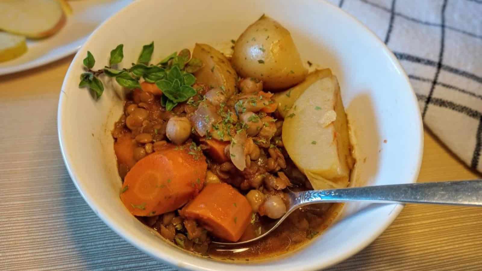 bowl of cholent with potatoes, carrots, lentils, chickpeas, and fresh thyme sprig and a spoon.