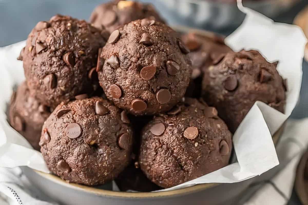 A bowl filled with chocolate muffins topped with chocolate chips, placed on a piece of parchment paper. The muffins appear moist and rich, showcasing a deep brown color.