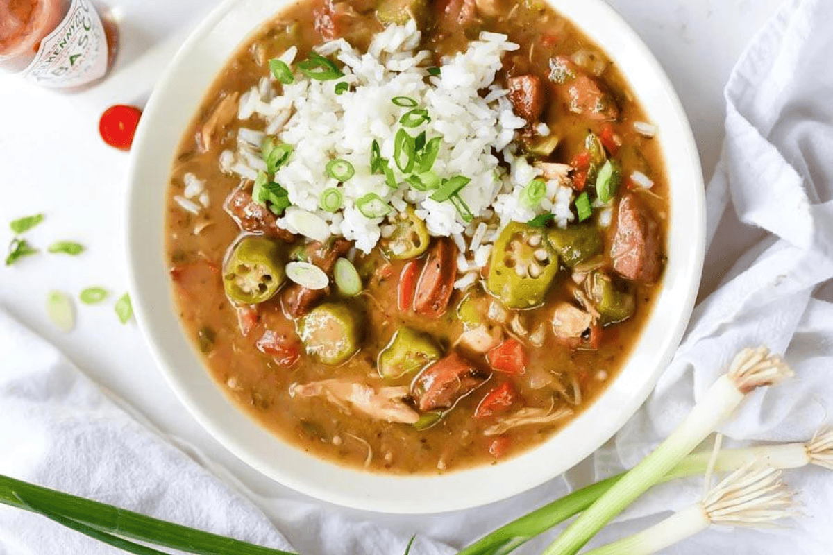 A bowl of gumbo topped with white rice and chopped green onions. The gumbo includes okra, diced tomatoes, sausage, and chicken. Surrounding the bowl are ingredients such as fresh green onions, a red chili, and a white napkin on a light-colored surface.