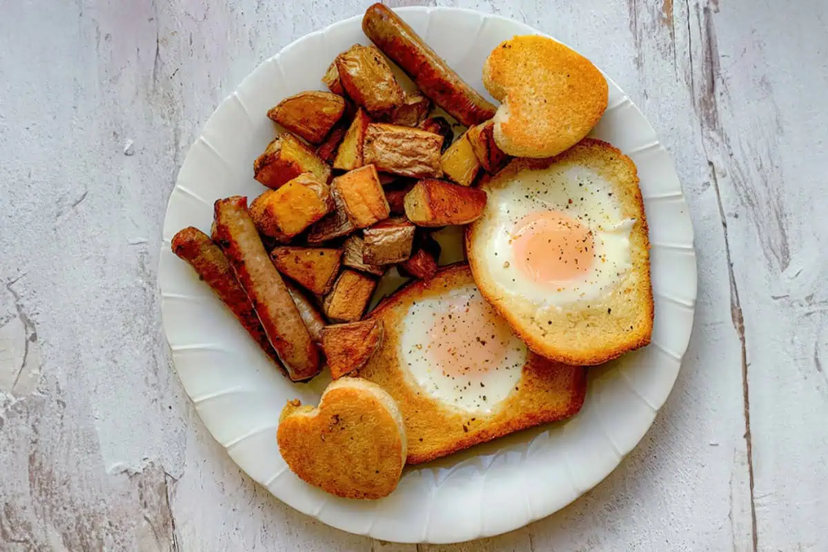 A white plate holds a breakfast of two sunny-side-up eggs cooked in toast cut into heart shapes, alongside diced roasted potatoes and several breakfast sausages. The wooden table beneath adds a rustic touch to the meal.
