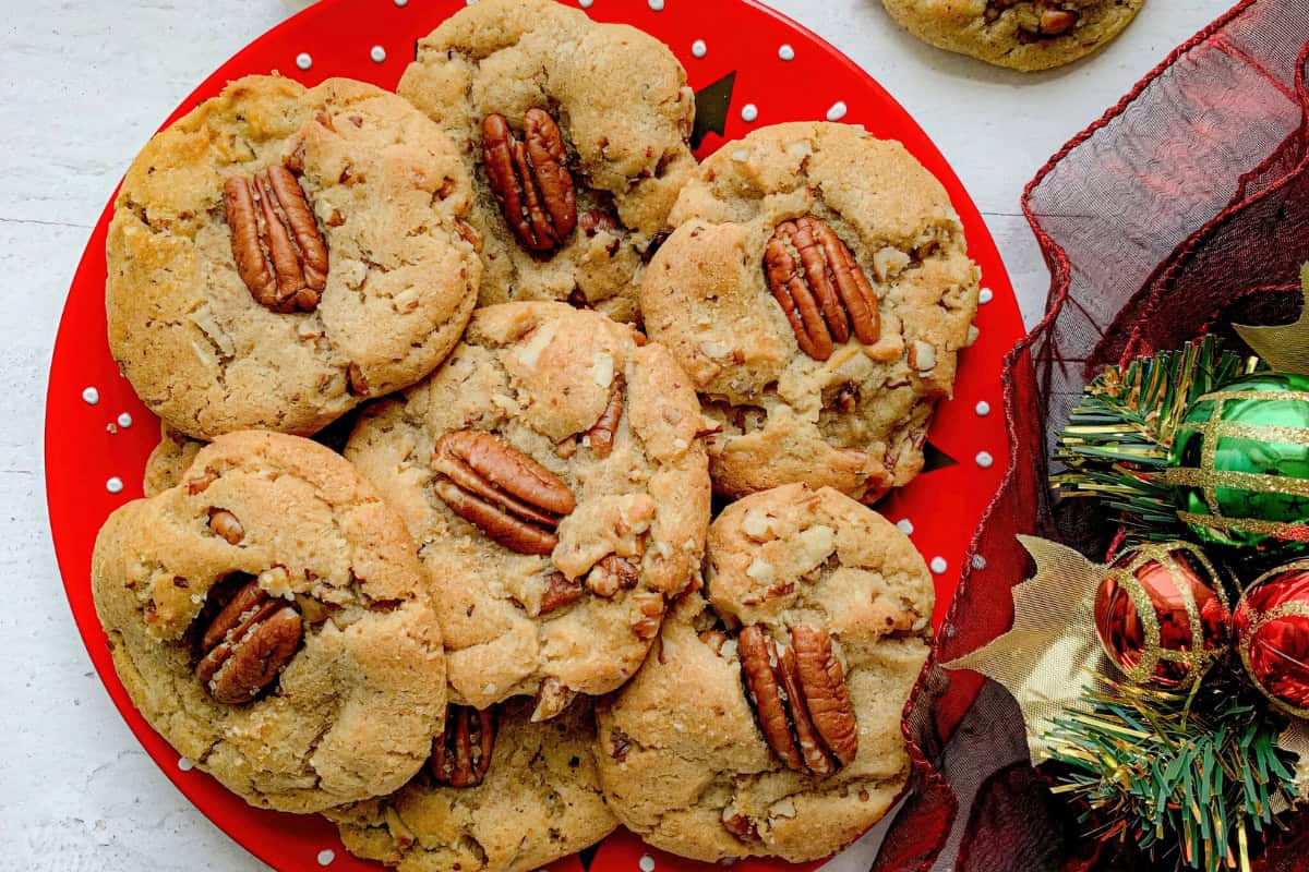 A red plate filled with homemade pecan cookies, each topped with a whole pecan. The plate is placed on a white surface, with some holiday decorations, including green and red ornaments and pine leaves, visible beside it.