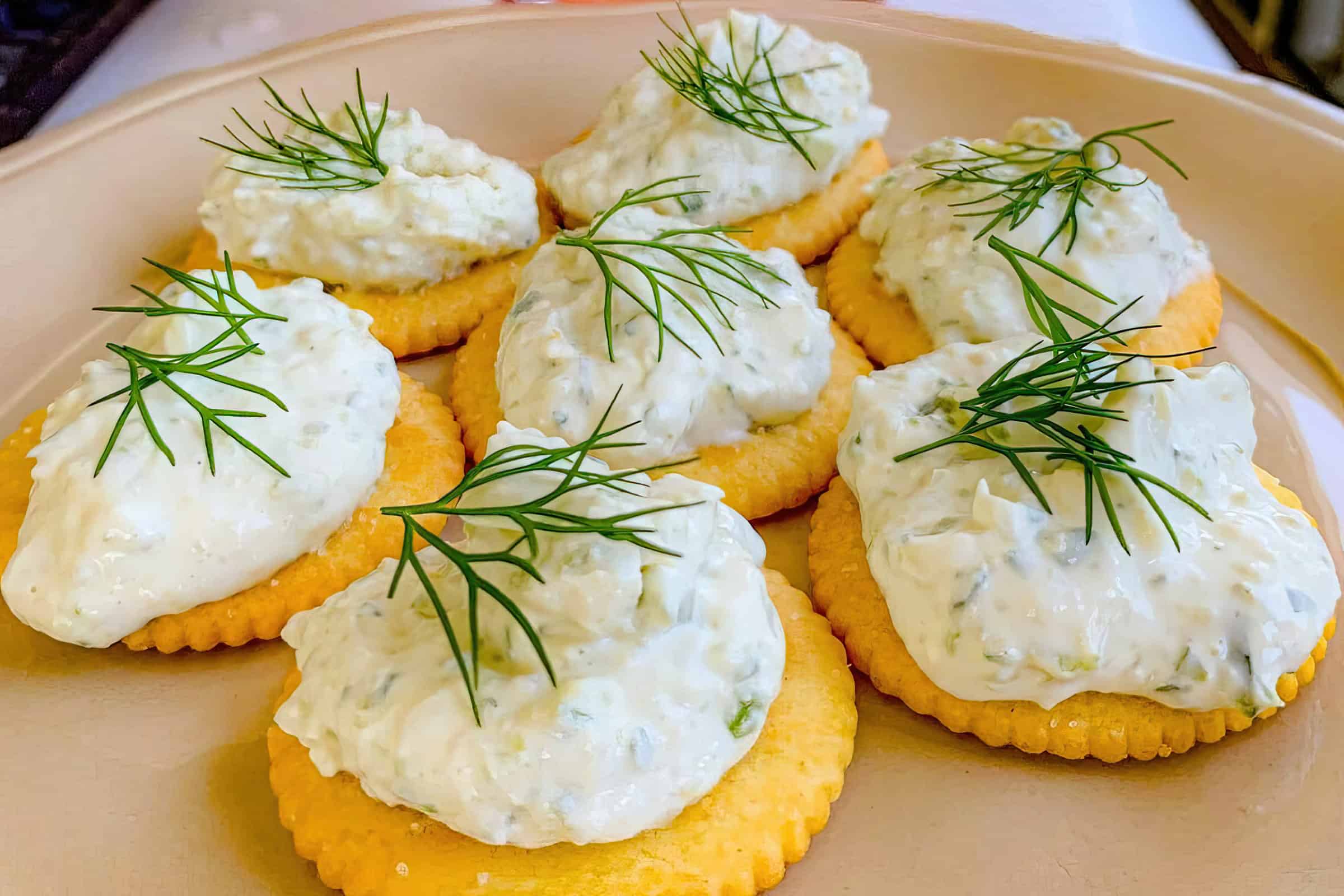 A plate of round crackers topped with generous dollops of creamy dip, garnished with small sprigs of fresh dill. The dish is presented on a beige plate, showcasing a delectable and appealing appetizer.