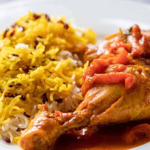 A plate features a serving of golden, fluffy saffron rice mixed with white rice and dotted with small red berries or spices. Beside the rice is a juicy, well-seasoned chicken leg covered in a rich, red tomato-based sauce. The dish appears appetizing and aromatic.