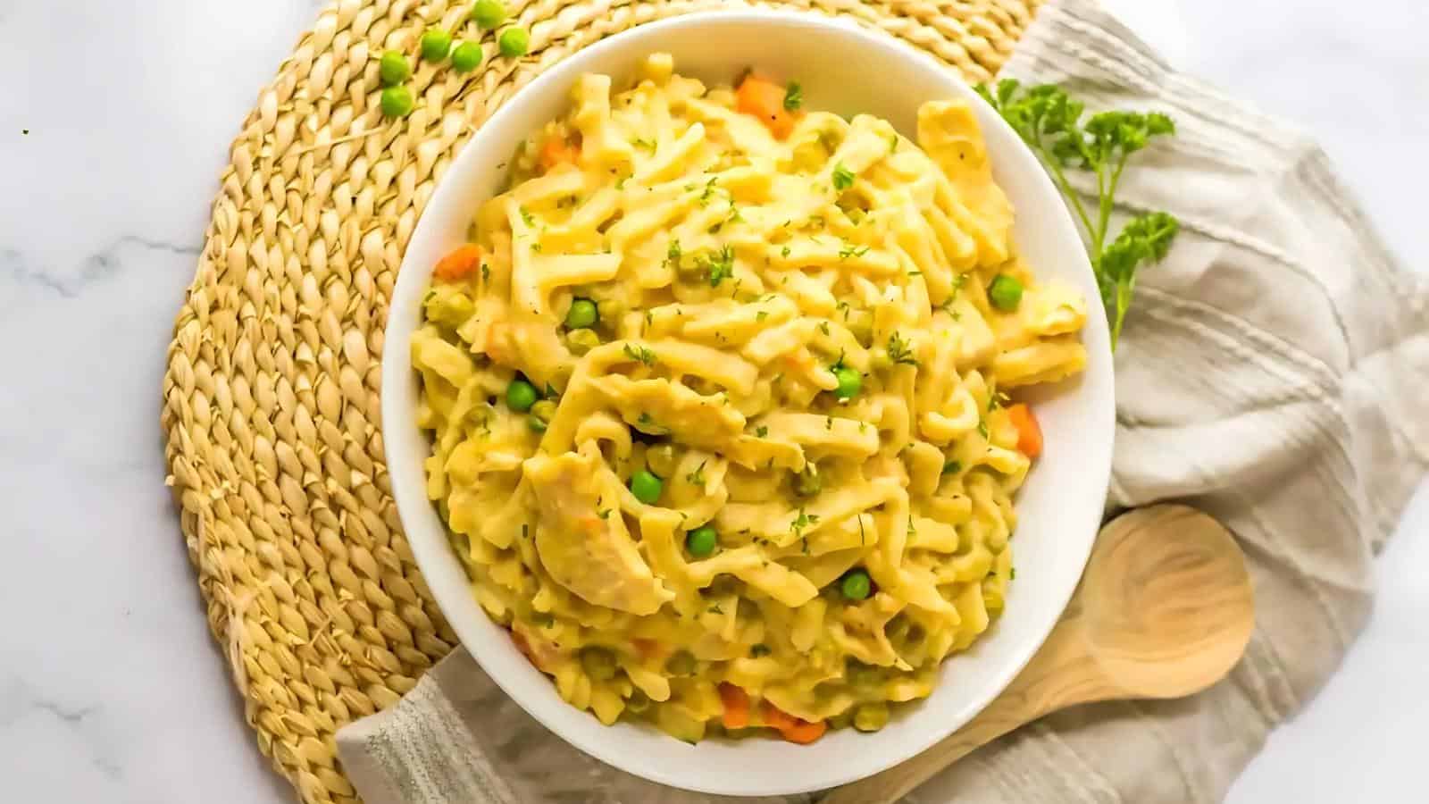 A bowl of creamy pasta with peas and carrots, a wooden spoon, and a cloth napkin on a woven placemat.