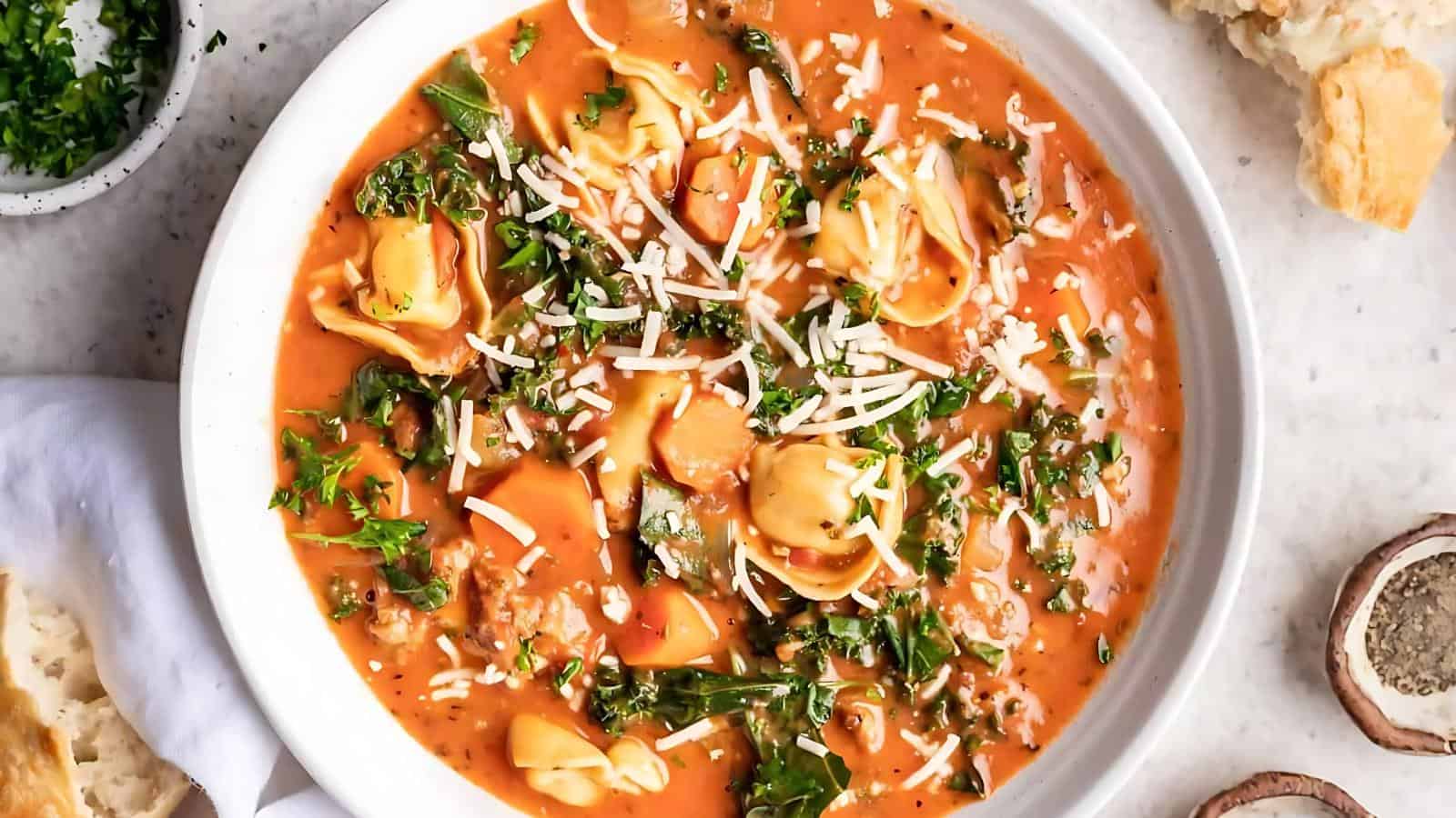 A bowl of tomato-based soup containing tortellini and greens, topped with cheese, with a side of bread.