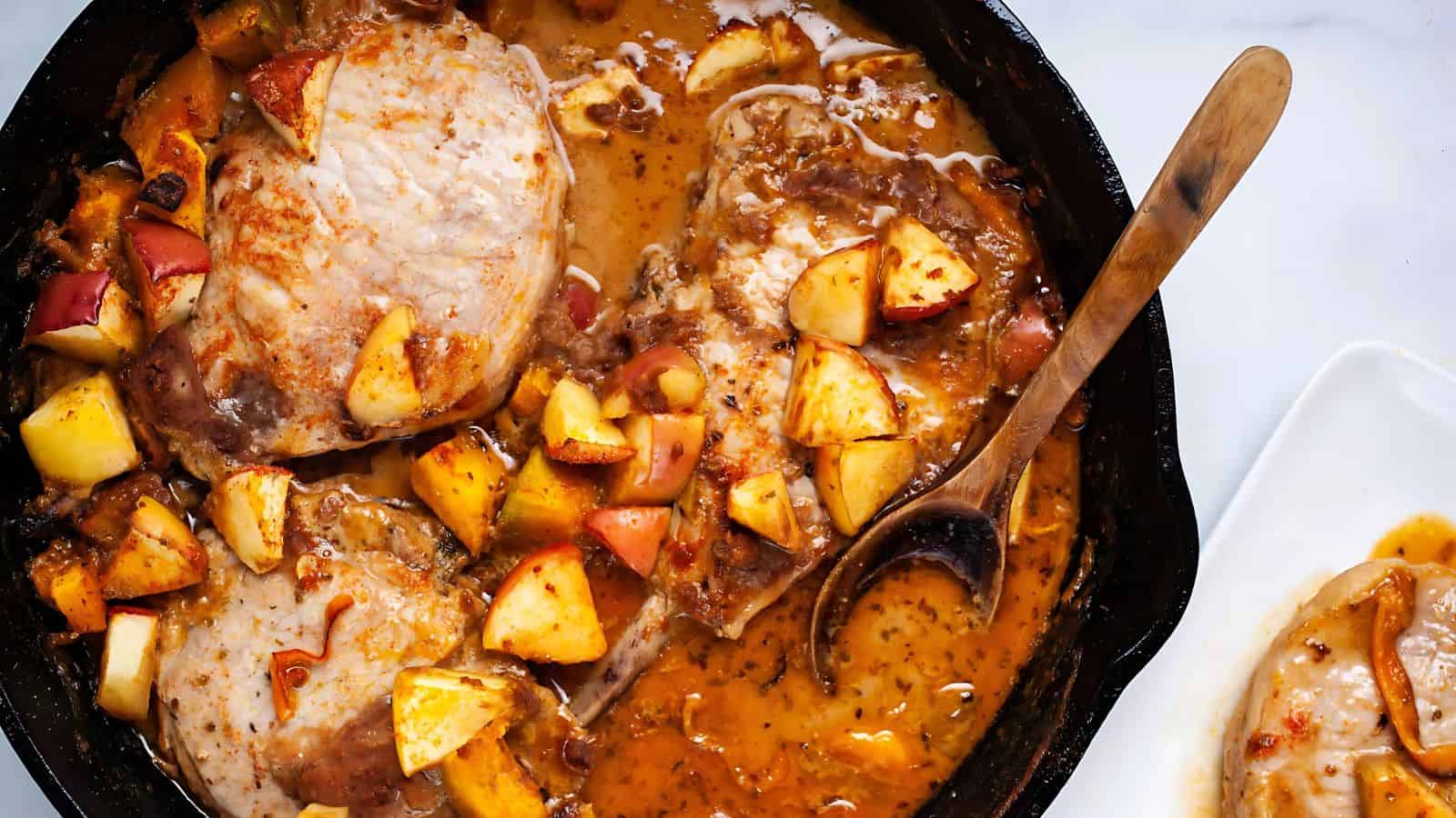 A skillet filled with pork chops cooked in a savory sauce, topped with diced apples and herbs. There's a wooden spoon resting in the skillet, and part of a white plate is visible on the side.