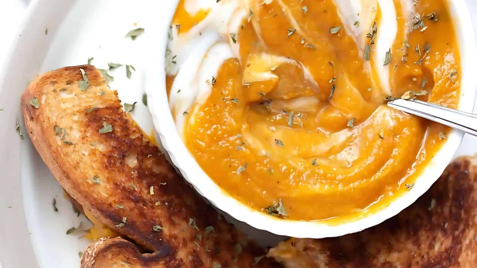 A bowl of creamy pumpkin soup with a swirl of cream on top, garnished with herbs. A spoon rests in the bowl. Two slices of toasted bread are on the side on a white plate.