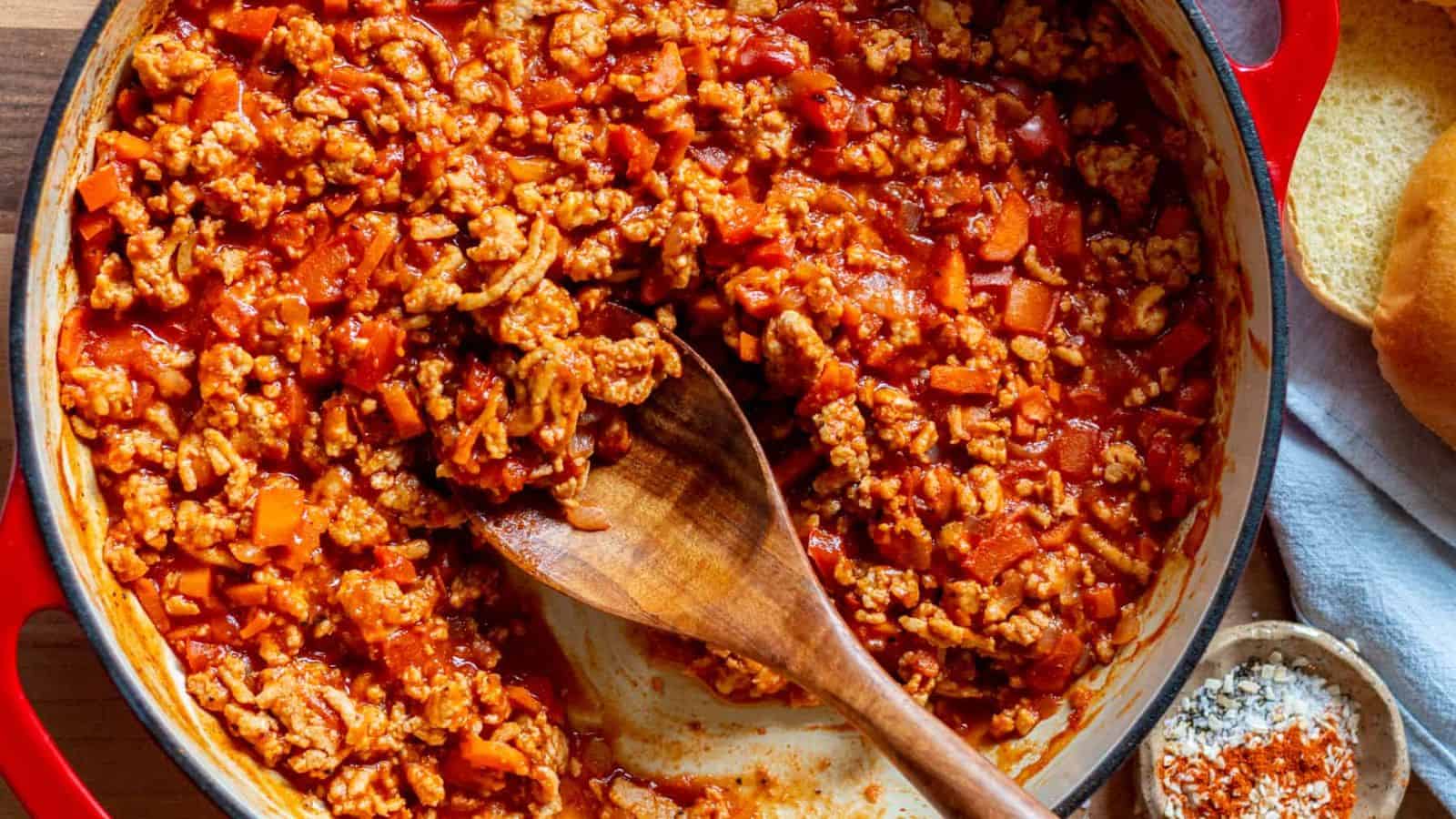 A red pot filled with cooked ground meat in tomato sauce, with visible chopped vegetables. A wooden spoon rests in the pot, and a small bowl of seasoning is nearby on a wooden surface.