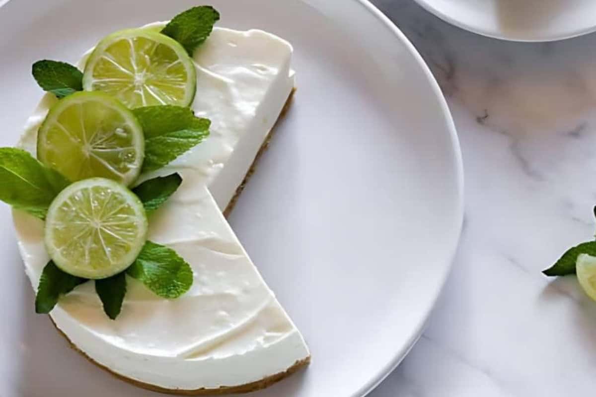 A slice of lime cheesecake topped with fresh lime slices and mint leaves sits on a white plate against a marble surface. The cheesecake has a creamy texture and a graham cracker crust.