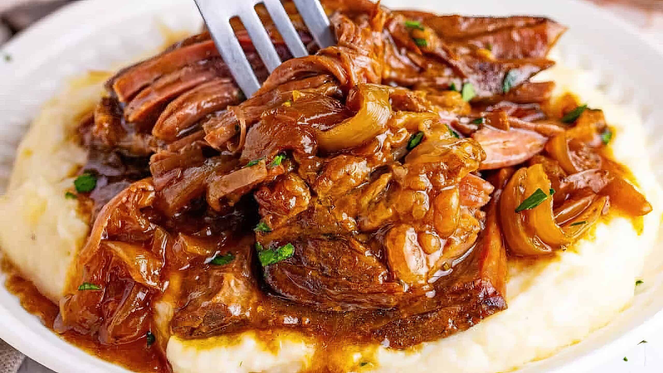 A plate of tender braised meat with caramelized onions served over creamy mashed potatoes. A fork is pulling apart the hearty meat on top, garnished with fresh herbs.