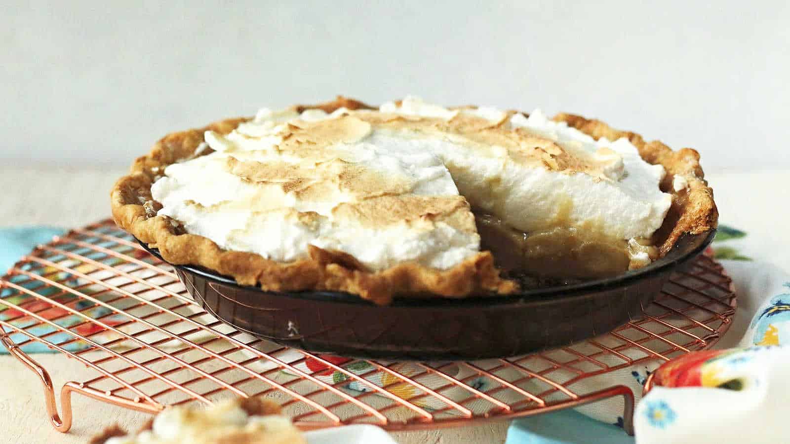 A freshly baked brown pie with a golden, flaky crust and a meringue topping is displayed on a copper cooling rack. A slice has been cut out, revealing the creamy filling inside. The pie is accompanied by a light blue cloth with a colorful floral pattern.