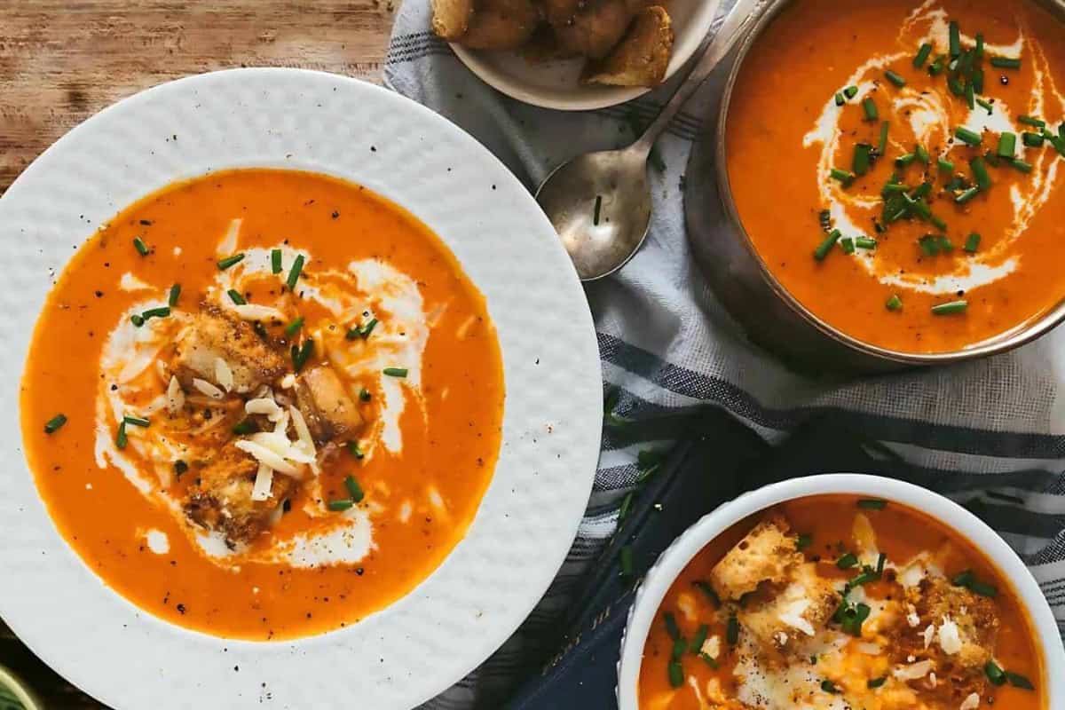 A table with bowls of creamy tomato soup garnished with bread croutons, chopped herbs, and a drizzle of cream. A spoon rests on the table along with a small bowl of bread pieces.