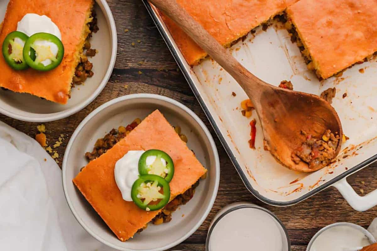 A casserole dish with a missing square of cornbread-topped chili, served on two plates. Each plate has a piece of the cornbread chili, topped with sour cream and sliced jalapeños. A wooden spoon rests in the dish, and two empty bowls are nearby.