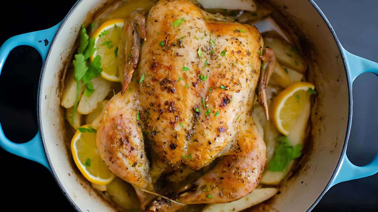 A roasted chicken seasoned with herbs is in a blue pot surrounded by lemon slices and potato wedges. The chicken is golden brown and garnished with parsley, creating a savory and inviting dish.