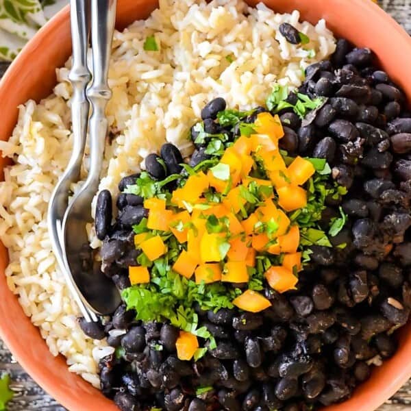 The image features a bowl filled with brown rice, topped with black beans, diced orange bell peppers, and chopped cilantro. Two spoons rest on the rice, and lime wedges are positioned beside the bowl.
