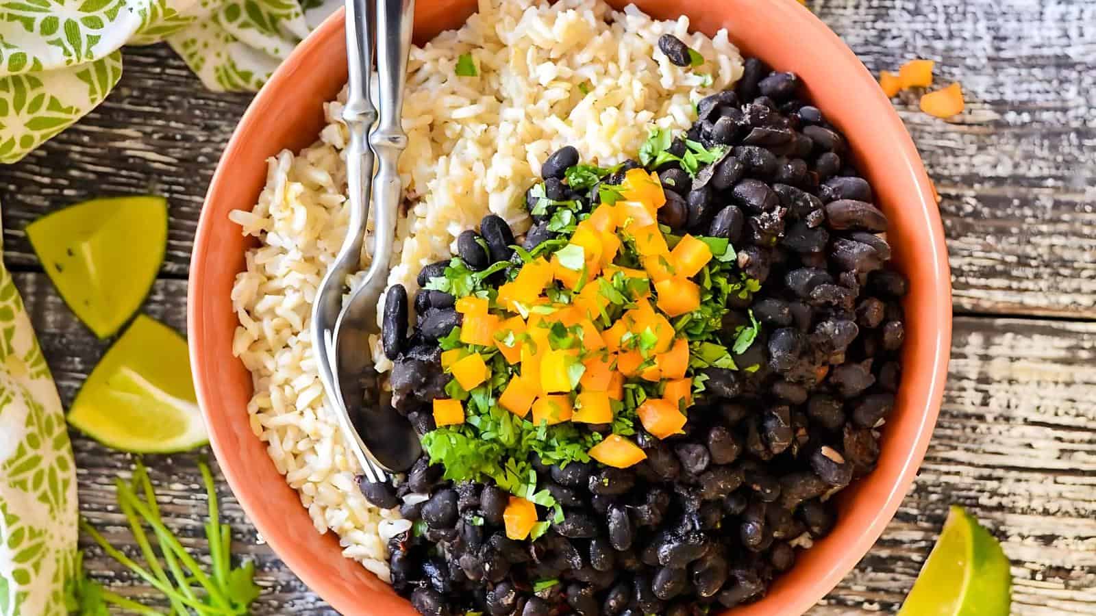 The image features a bowl filled with brown rice, topped with black beans, diced orange bell peppers, and chopped cilantro. Two spoons rest on the rice, and lime wedges are positioned beside the bowl.
