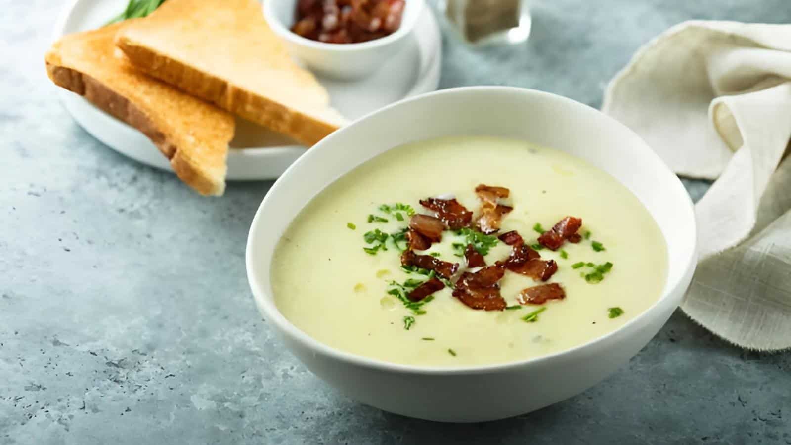 A bowl of creamy soup topped with crispy bacon and herbs is in the foreground, accompanied by toast and a small dish of bacon bits in the background on a gray surface.