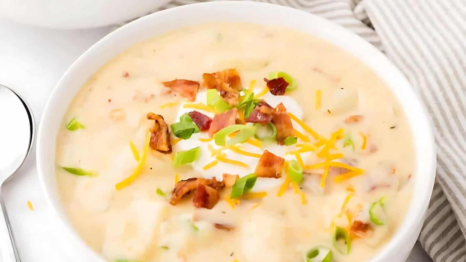 A bowl of creamy soup with bacon bits, shredded cheese, and chopped green onions beside a striped cloth napkin.
