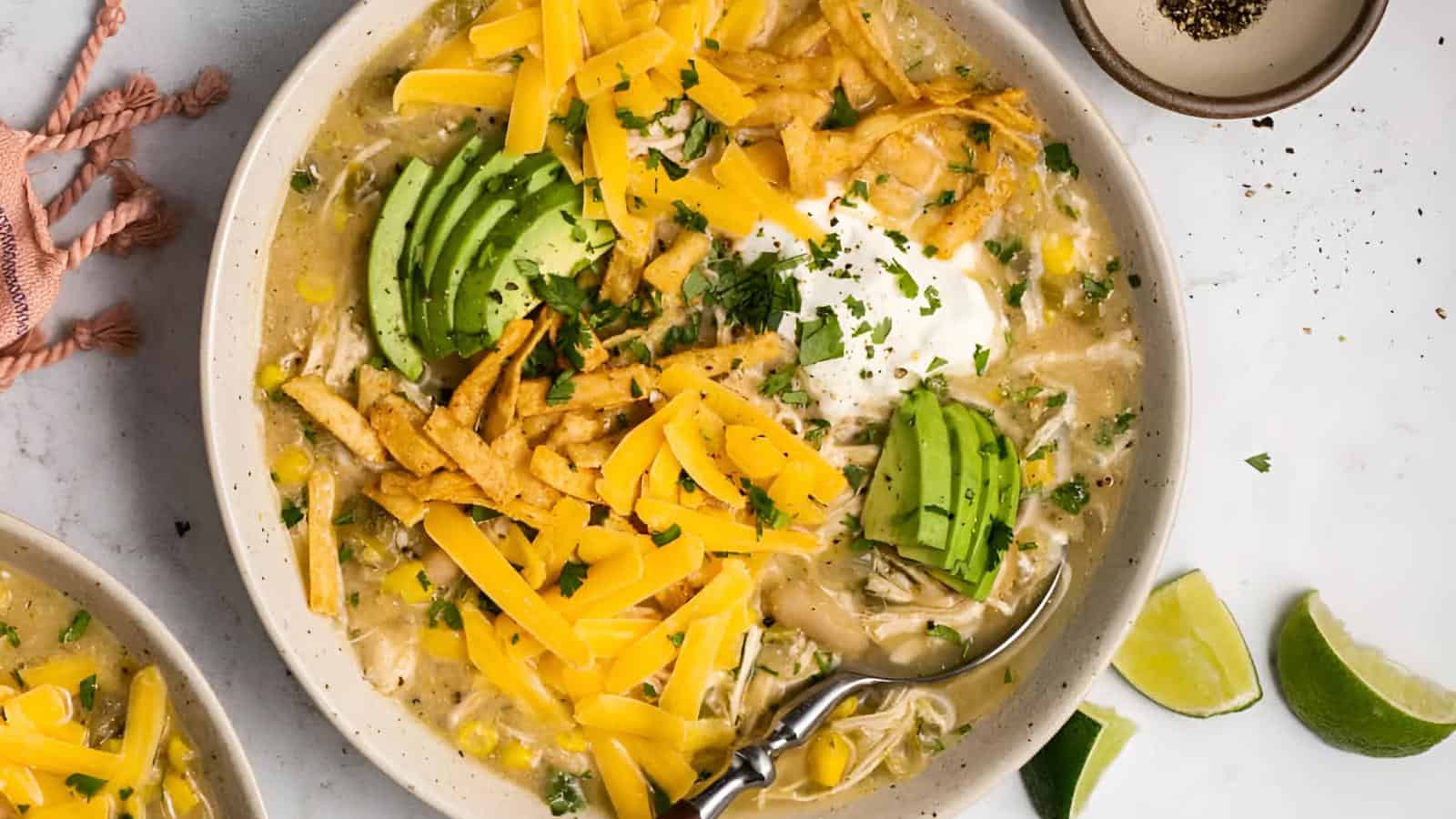 A bowl of chicken tortilla soup topped with sliced avocado, shredded cheese, fried tortilla strips, and a dollop of sour cream. Garnished with chopped herbs. A spoon rests in the bowl. Lime wedges and a bowl of pepper are beside it.