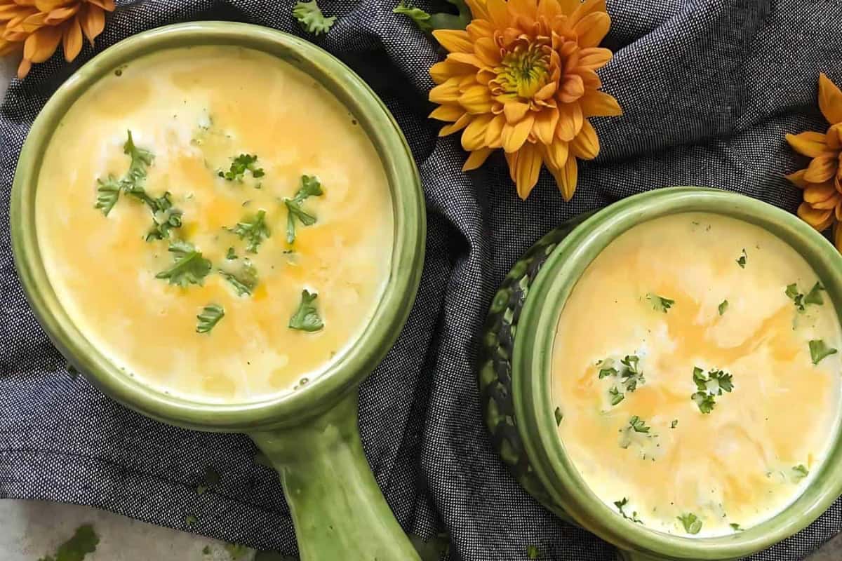 Two green ceramic bowls filled with creamy soup, garnished with chopped herbs. They are placed on a dark gray cloth, surrounded by vibrant orange flowers.