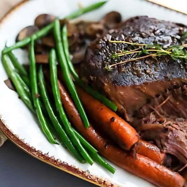 A plate featuring roast beef covered in gravy, green beans, carrots, mushrooms, and a garnish of herbs. A fork is positioned beside the plate.