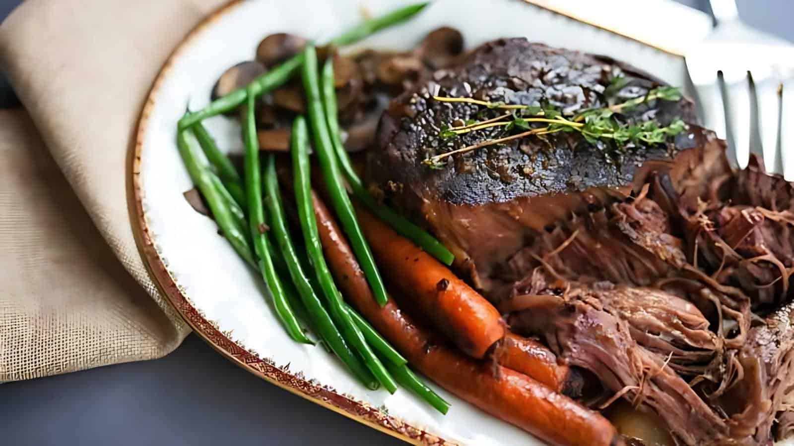 A plate featuring roast beef covered in gravy, green beans, carrots, mushrooms, and a garnish of herbs. A fork is positioned beside the plate.