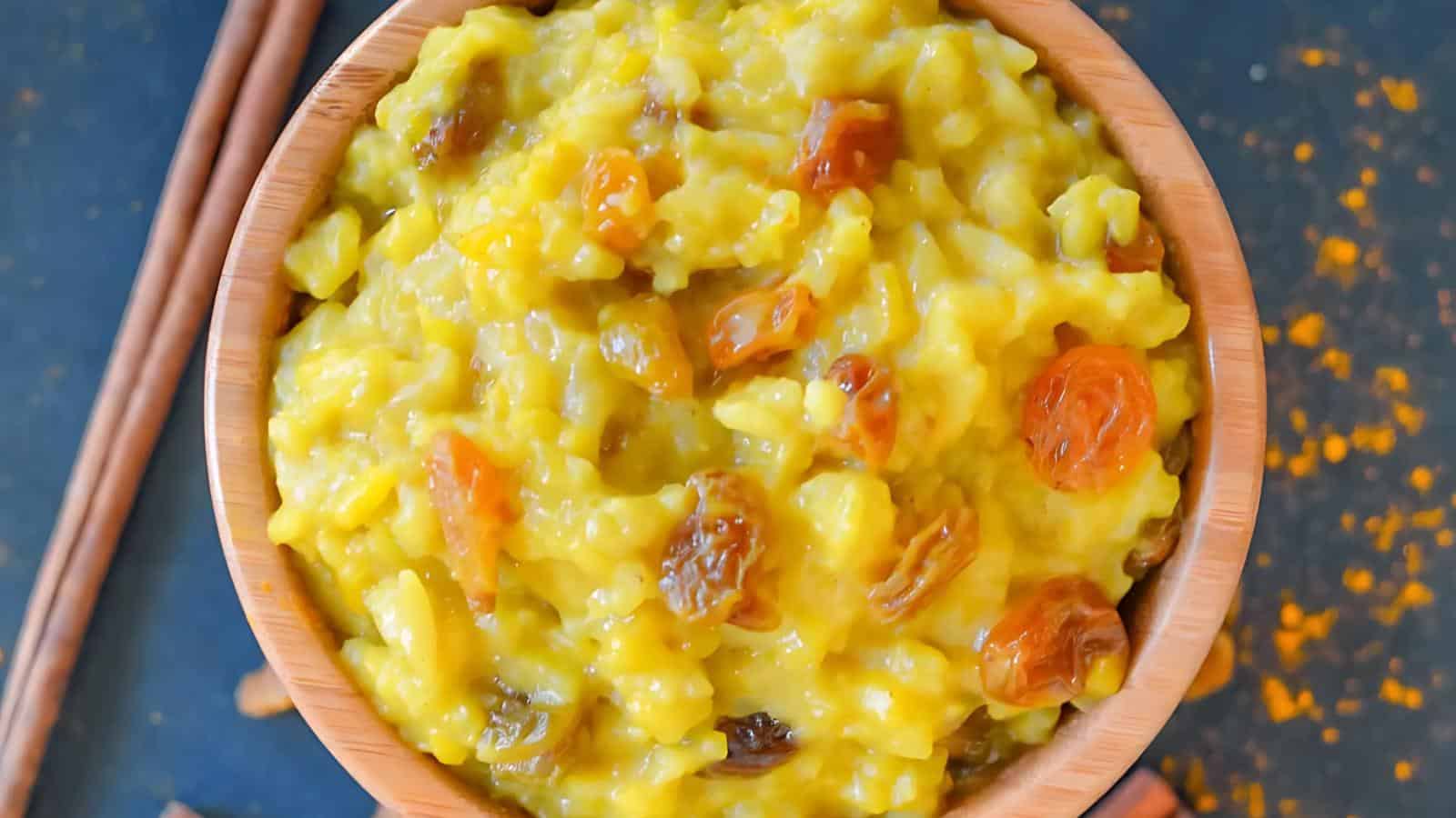 A bowl of yellow-colored rice pudding topped with raisins on a dark surface.