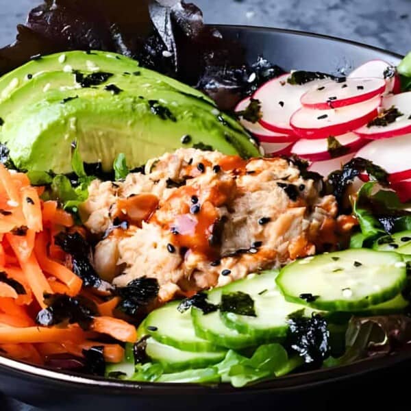 A bowl with sliced avocado, radishes, carrots, cucumber, greens, and mashed tuna topped with sesame seeds and seaweed.