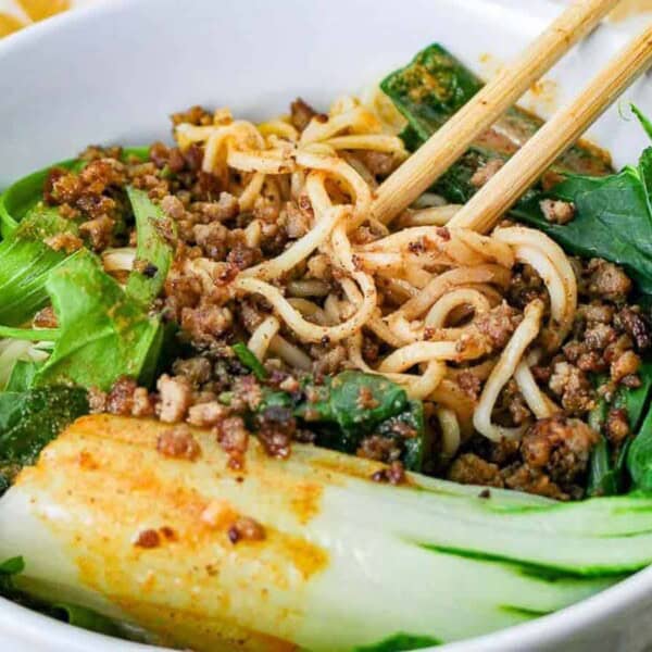 A white bowl with noodles, ground meat, bok choy, leafy greens, and chopsticks resting on top.