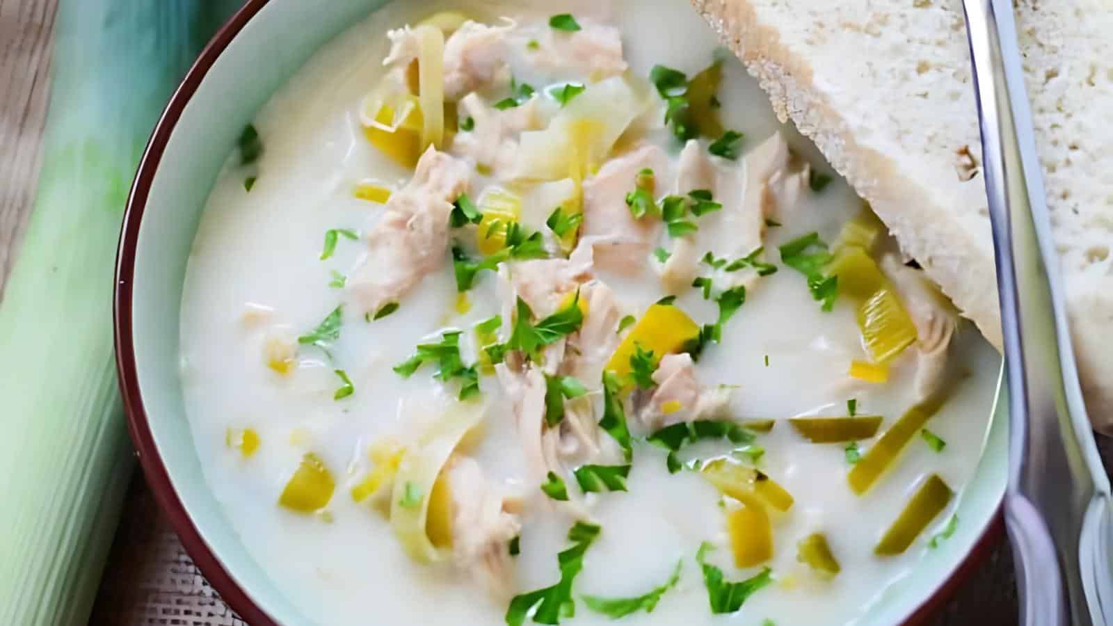 Bowl of creamy soup with chunks of chicken and leek, topped with chopped herbs, alongside a slice of bread.