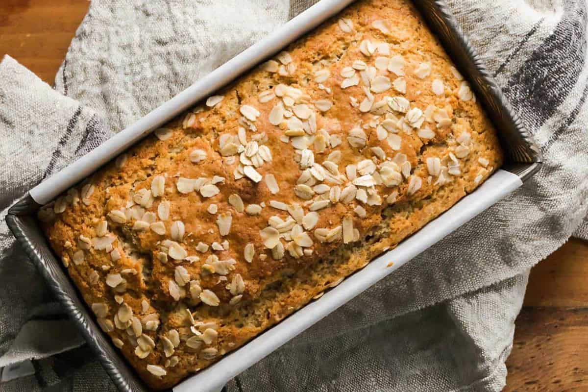 A freshly baked loaf of bread in a rectangular pan, topped with oats. The pan rests on a light-colored cloth, creating a warm and rustic presentation.