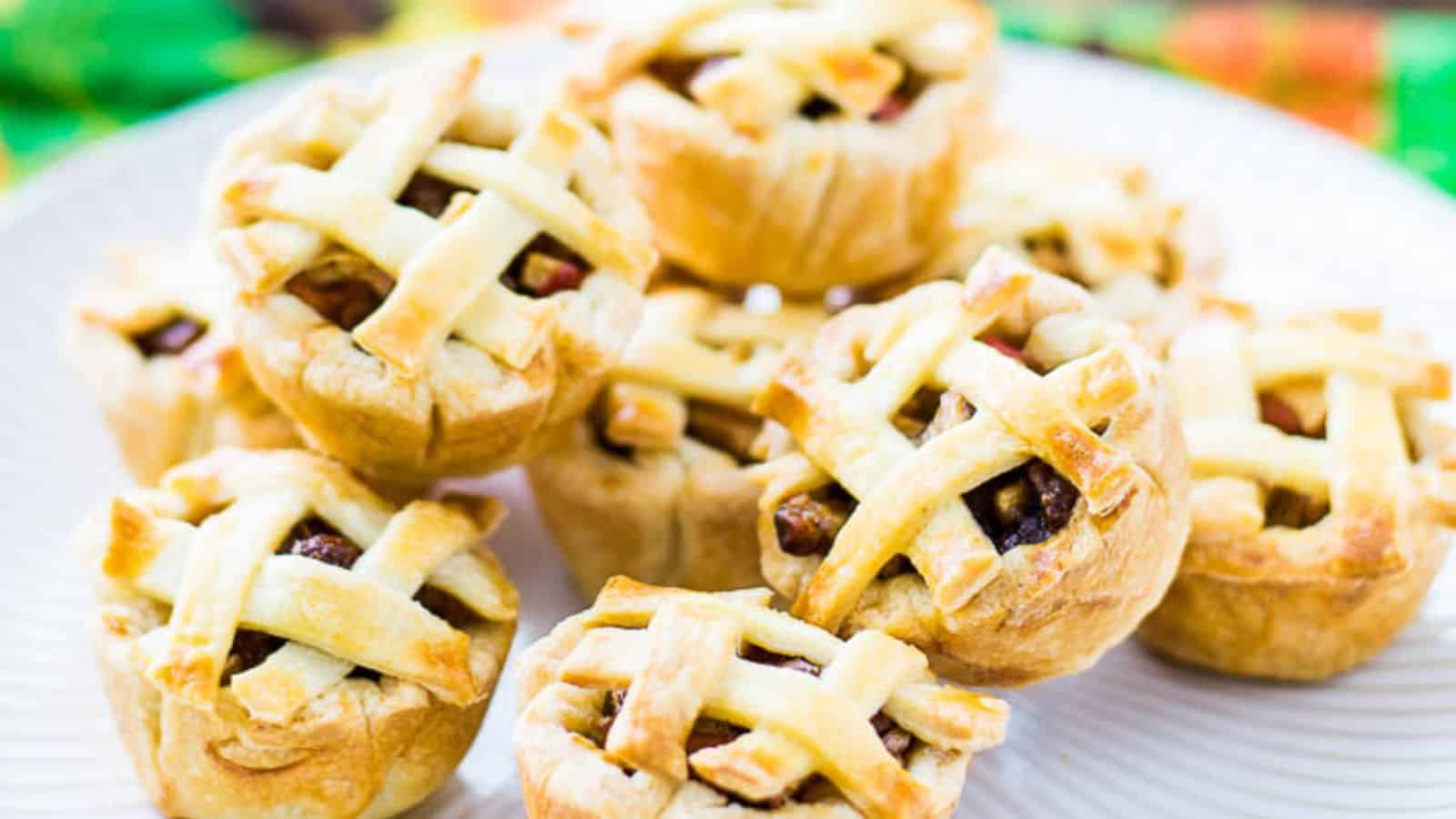 A close-up shot of a plate filled with mini apple pies featuring a lattice crust. The pies are golden brown, and the filling is visible through the lattice. The background has a blurred, colorful pattern.