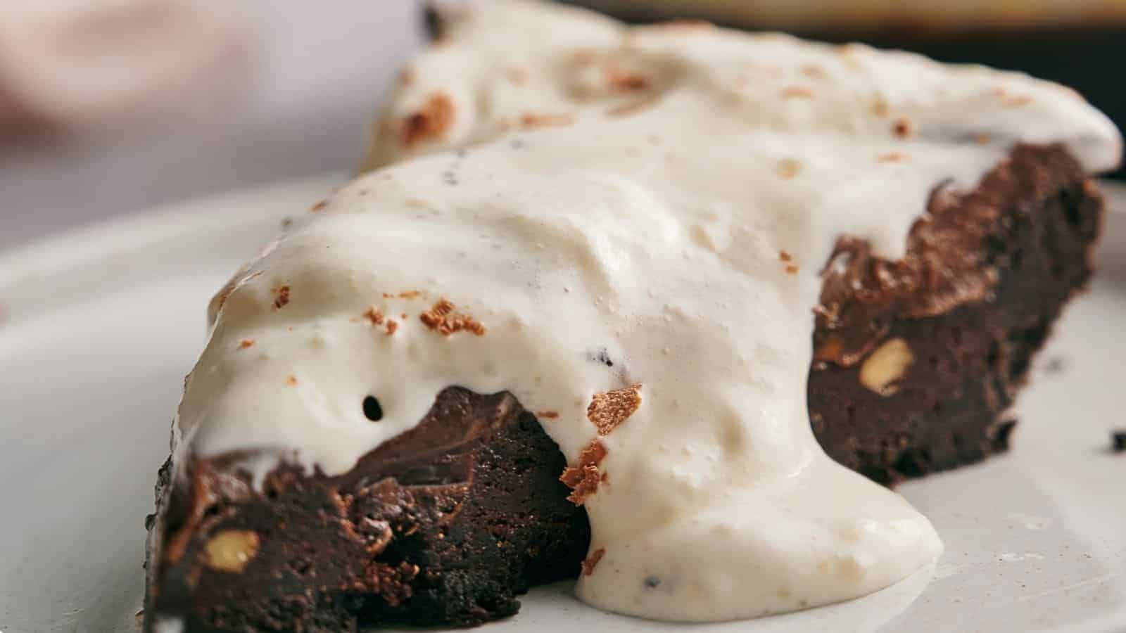 A slice of dense, dark chocolate brownie topped with a generous layer of creamy, white sauce. The brownie has visible nut pieces and a slightly crumbly texture, placed on a white plate.