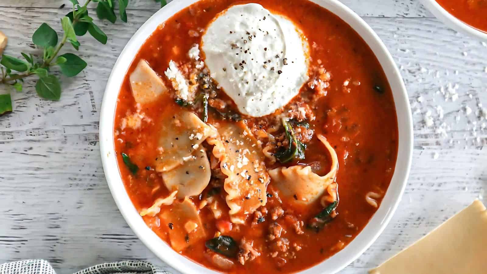 A bowl of lasagna soup garnished with a dollop of ricotta cheese and black pepper. The soup contains pasta, ground meat, and herbs, all in a rich tomato sauce. Fresh herbs are sprinkled on top and a plant is visible on the side.