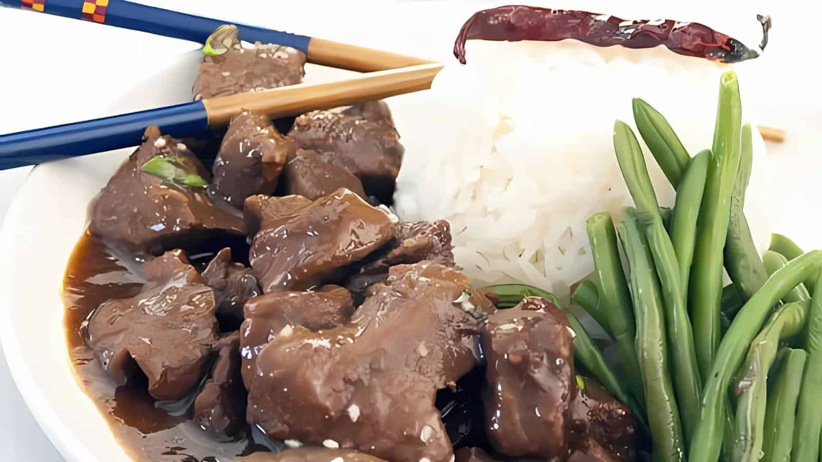 A plate with beef in sauce, accompanied by steamed white rice, green beans, a dried chili, and a pair of chopsticks.