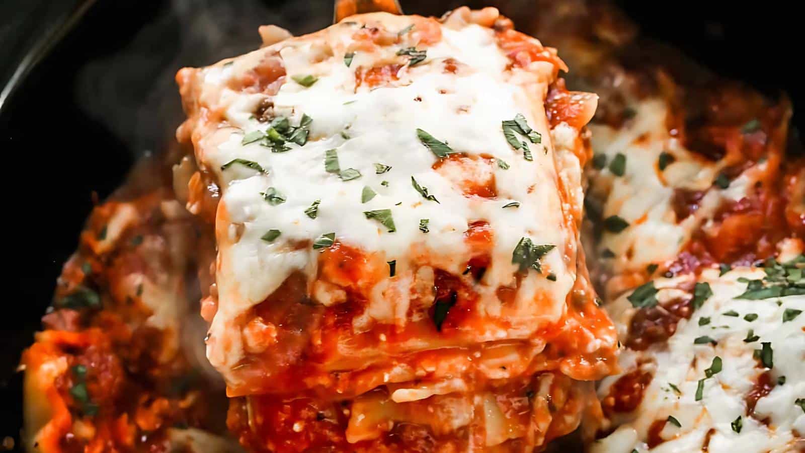 A close-up image of a lasagna slice featuring melted cheese, visible steam, and a sprinkling of herbs on top.