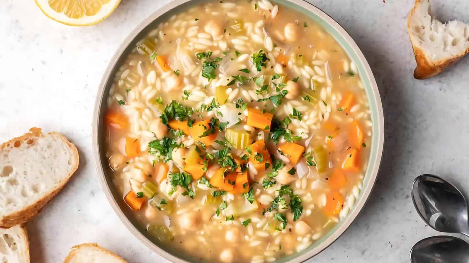 The image shows a bowl of soup containing orzo, diced carrots, celery, chickpeas, and a parsley garnish. Beside the bowl are slices of bread and a lemon wedge.
