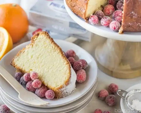 A slice of pound cake on a plate with sugared cranberries, an orange, and a cake stand holding more cake and cranberries behind.