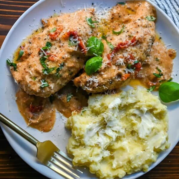 A plate with two pieces of chicken in creamy sauce, topped with herbs and cheese, served with mashed potatoes; fork and napkin nearby.