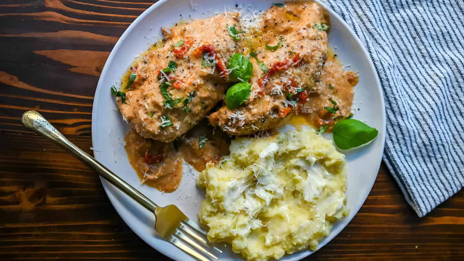 A plate with creamy chicken topped with chopped herbs and tomatoes, accompanied by mashed potatoes. A gold fork rests beside the food on a white plate, placed on a dark wooden table with a striped cloth nearby.