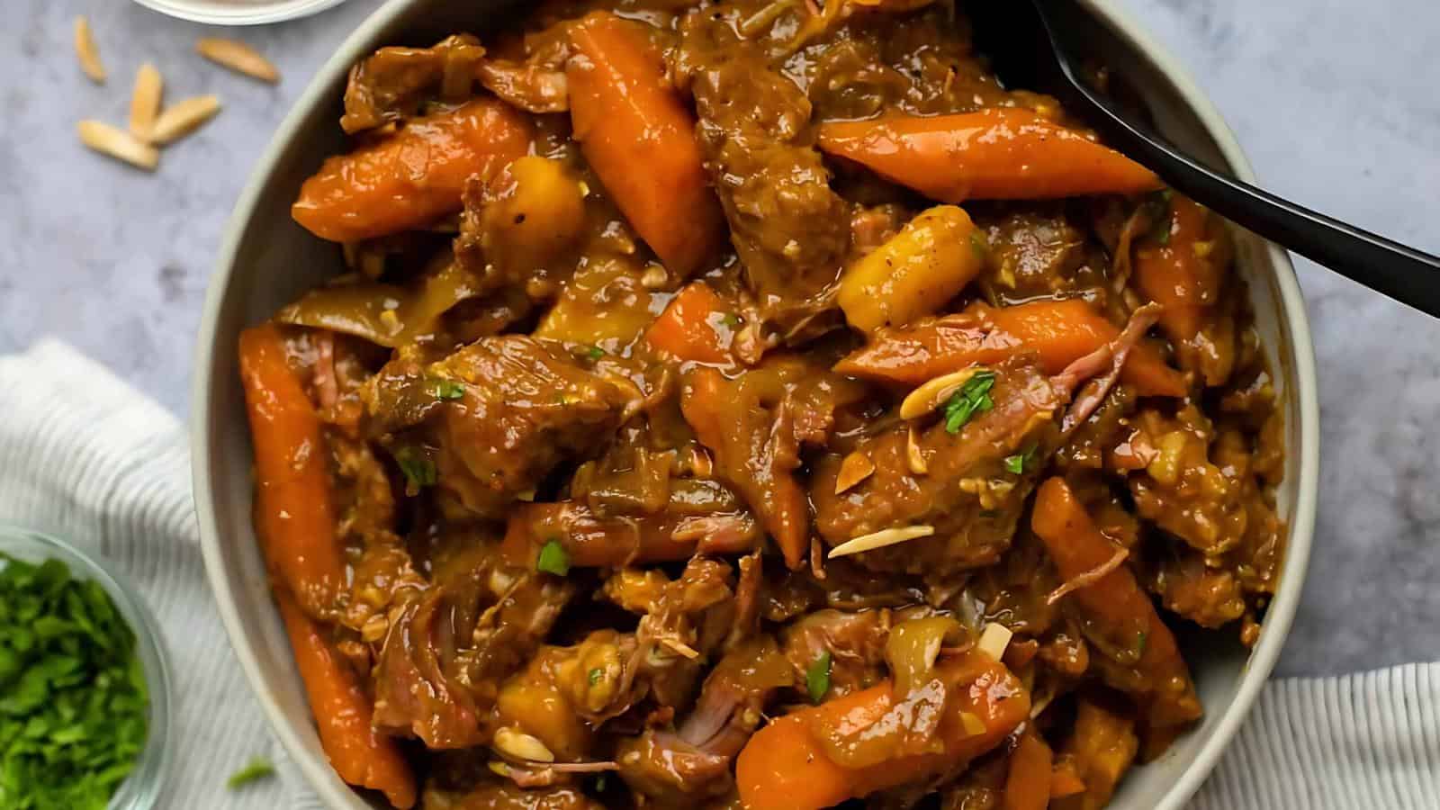 A bowl of beef stew with carrots and chunks of potatoes, garnished with herbs. A black spoon is in the bowl.