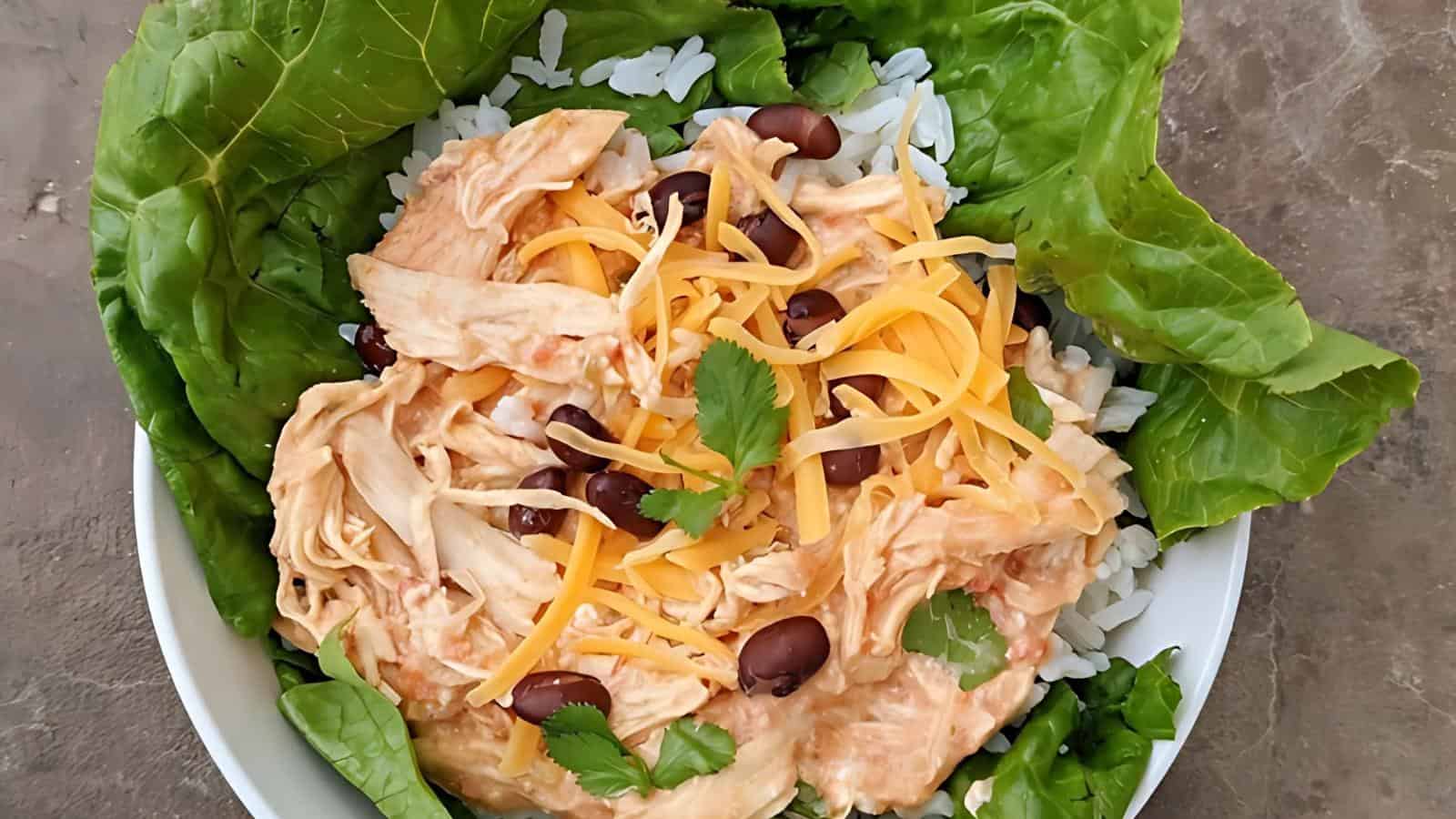 A white bowl containing rice, shredded chicken, black beans, shredded cheese, and garnished with cilantro. It is surrounded by green lettuce leaves.