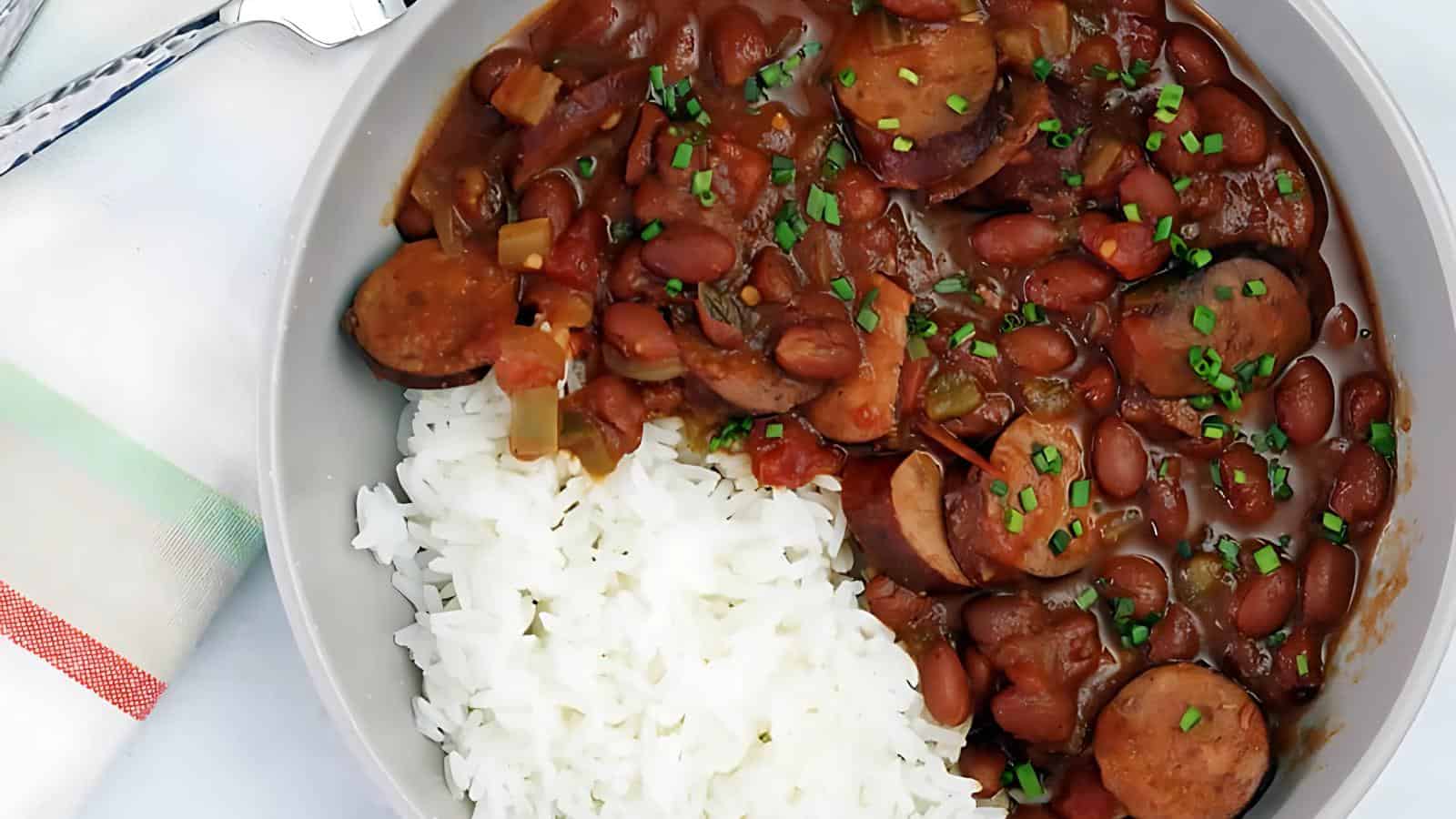 A bowl containing white rice mixed with red beans and sliced sausage, topped with green herbs. A fork lies on a striped cloth napkin next to the bowl.